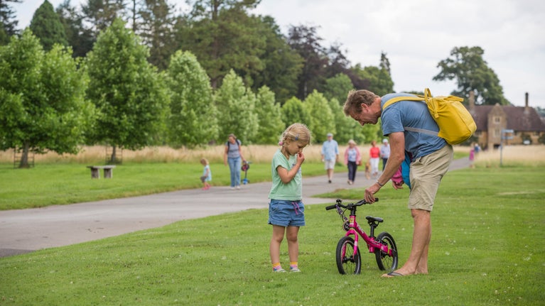 A girl looking at a balance bike with a parent holding it up.